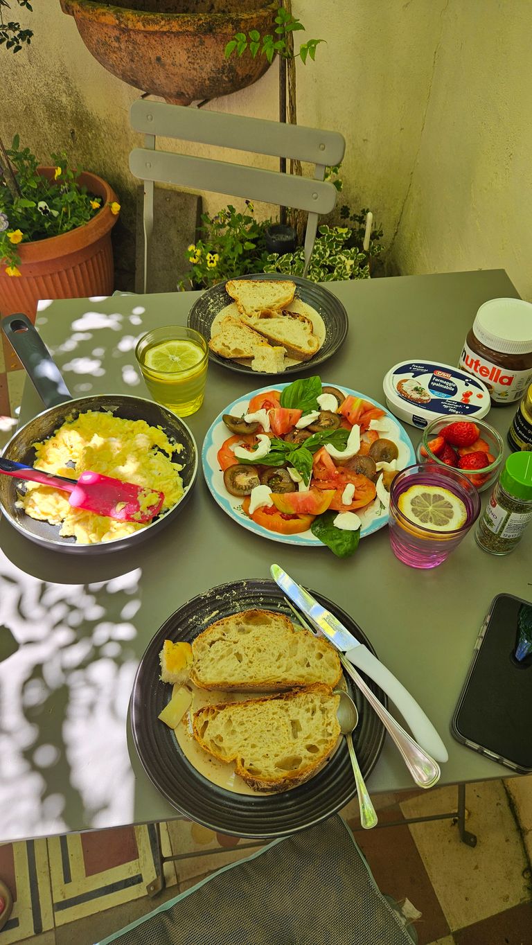 Tisch auf einer Terrasse mit Weißbrotscheiben auf Tellern, einem Teller mit Tomaten, Pilzen, Mozzarella und Basilikum, Rührei in einer Pfanne, einem Glas mit Zitronensaft, einem Glas mit roten Beeren und verschiedenen Aufstrichen darunter Nutella.