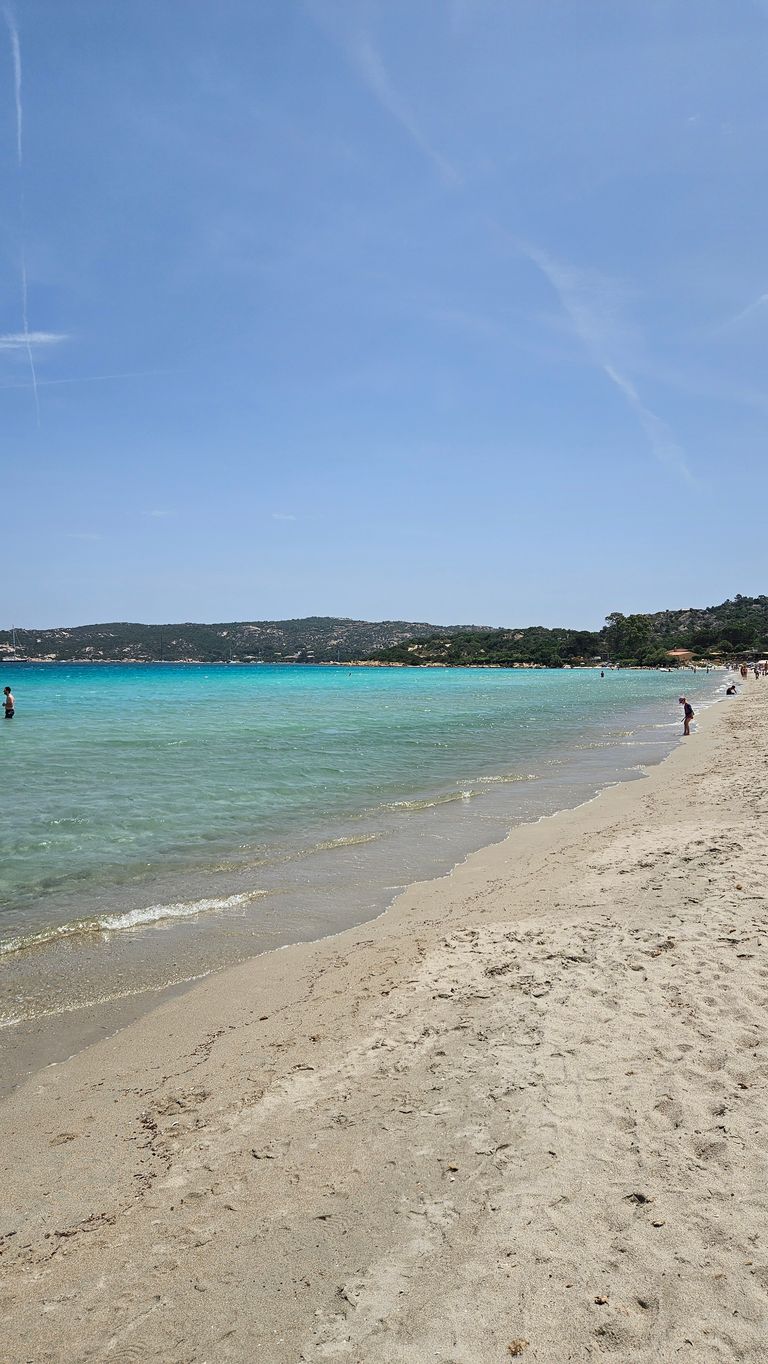 Ein weiter Sandstrand mit klarem, türkisfarbenem Wasser und vereinzelt badenden Menschen bei klarem Himmel. Im Hintergrund sind bewaldete Hügel zu sehen.
