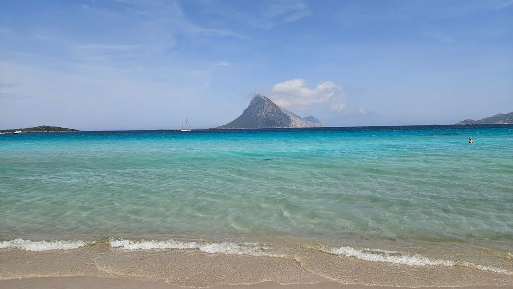 Klares türkisfarbenes Meer mit Sandstrand im Vordergrund und einer Insel mit bergigem Gipfel am Horizont unter blauem Himmel.