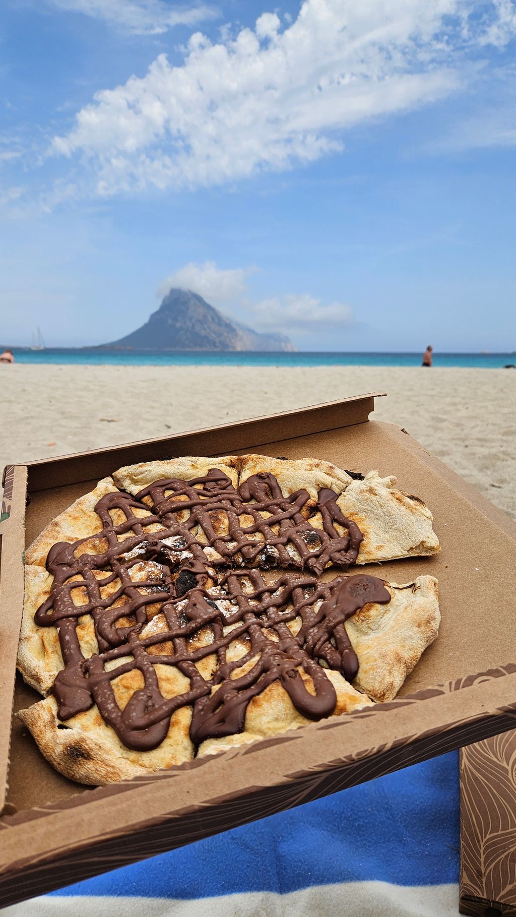 Ein Schokopizza in einer Pizzaschachtel auf einem Strand mit Sand, Meer und Berg im Hintergrund unter bewölktem Himmel.
