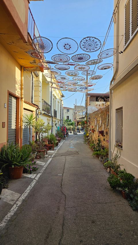 Narrow street with yellow and cream-colored buildings, round crocheted decorations hanging overhead, numerous potted plants along the street.