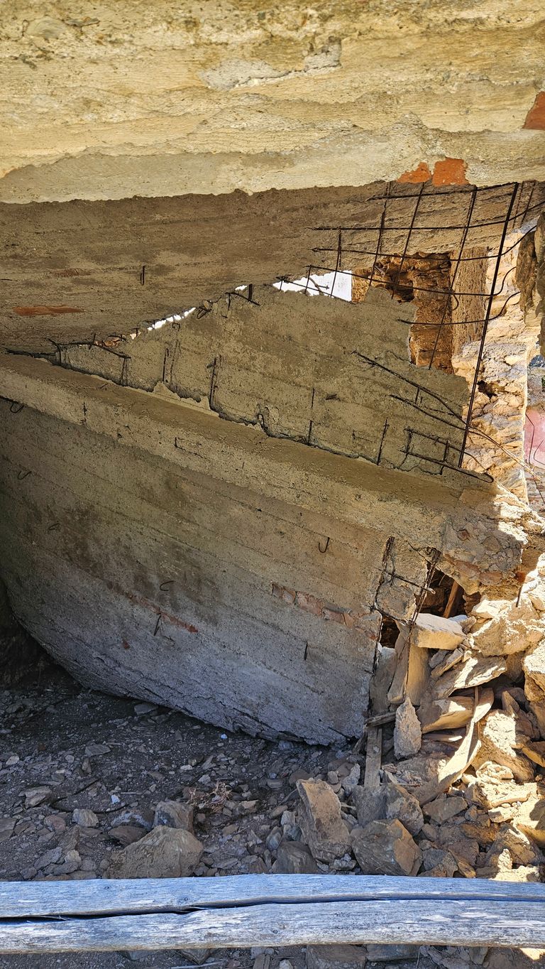 Riesiger Betonbrocken mit freiliegendem Stahlgerüst liegt auf unebenem felsigem Boden vor einer verfallenen Wand in einer Ruine.