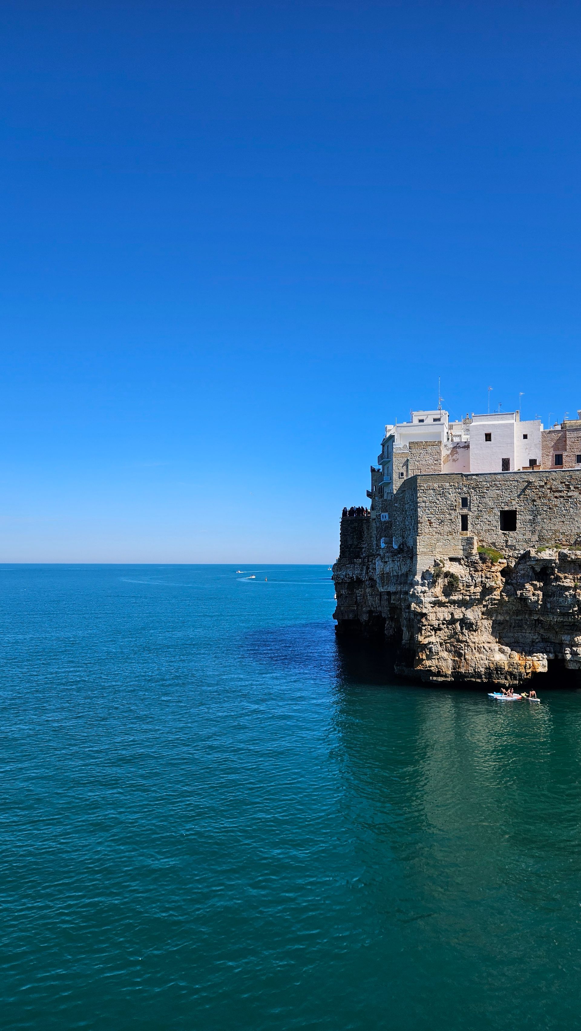 Eine steinerne Küstenstadt mit weißen Häusern, die auf hohen Klippen über einem ruhigen blauen Meer thront, unter klarem, wolkenlosem Himmel.